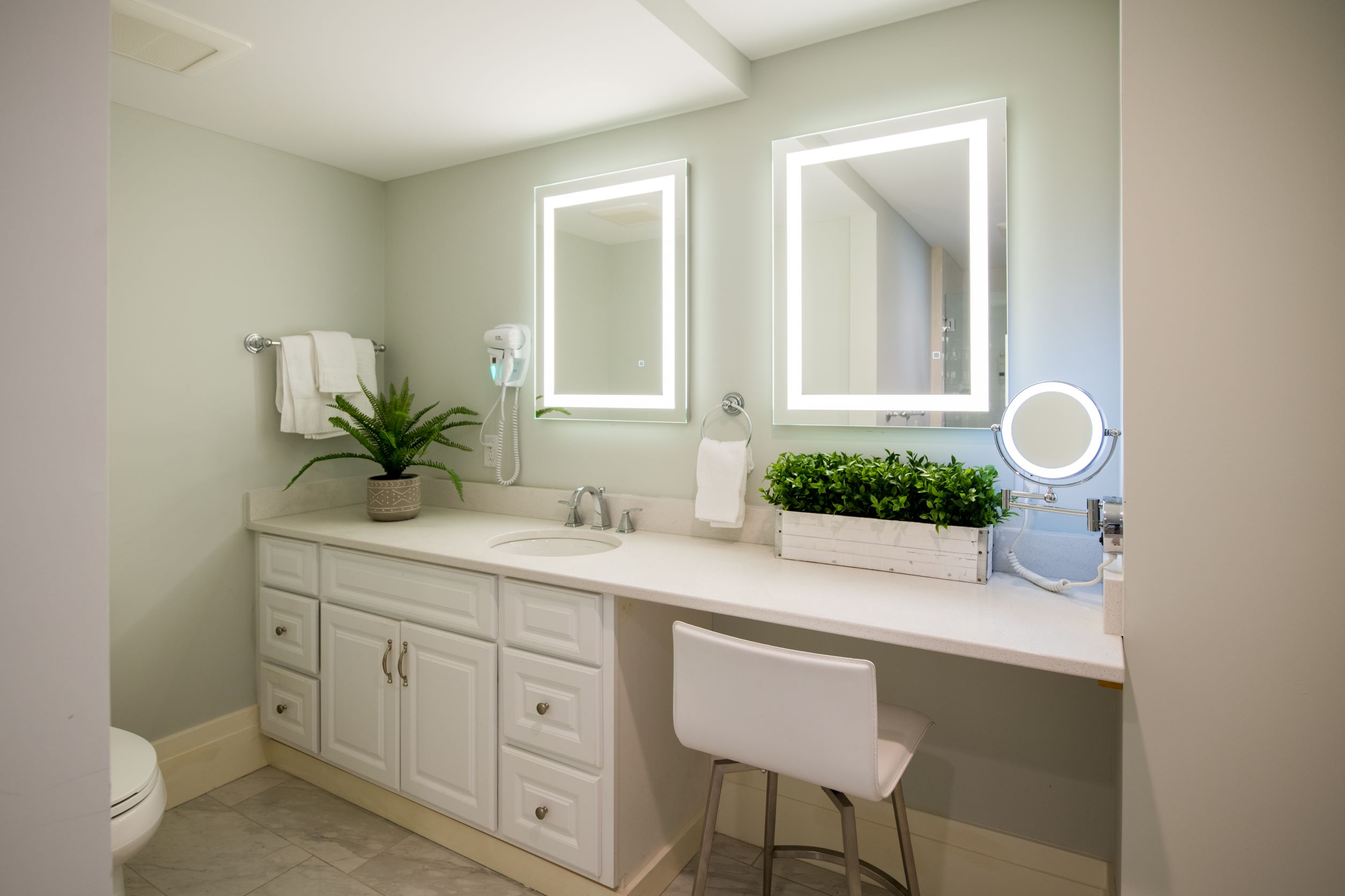 a bathroom vanity with sink and cabinets on one side, a white chair, and two lighted mirrors.