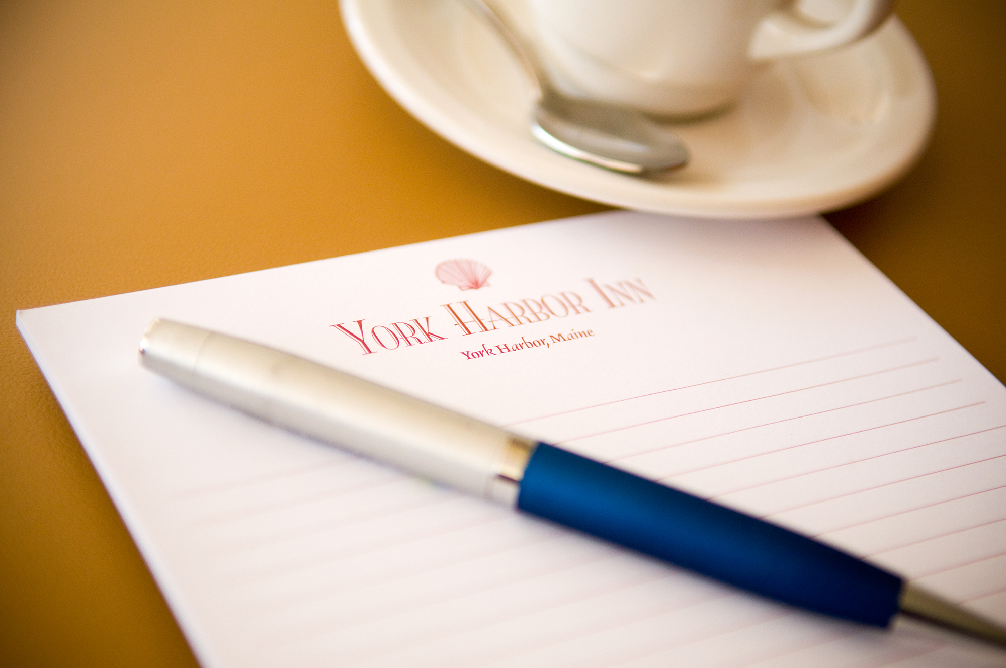 A blue and silver pen lays on top of a lined pad of a paper with a red logo on top of the paper. A coffee cup, saucer, and spoon sit in the background.