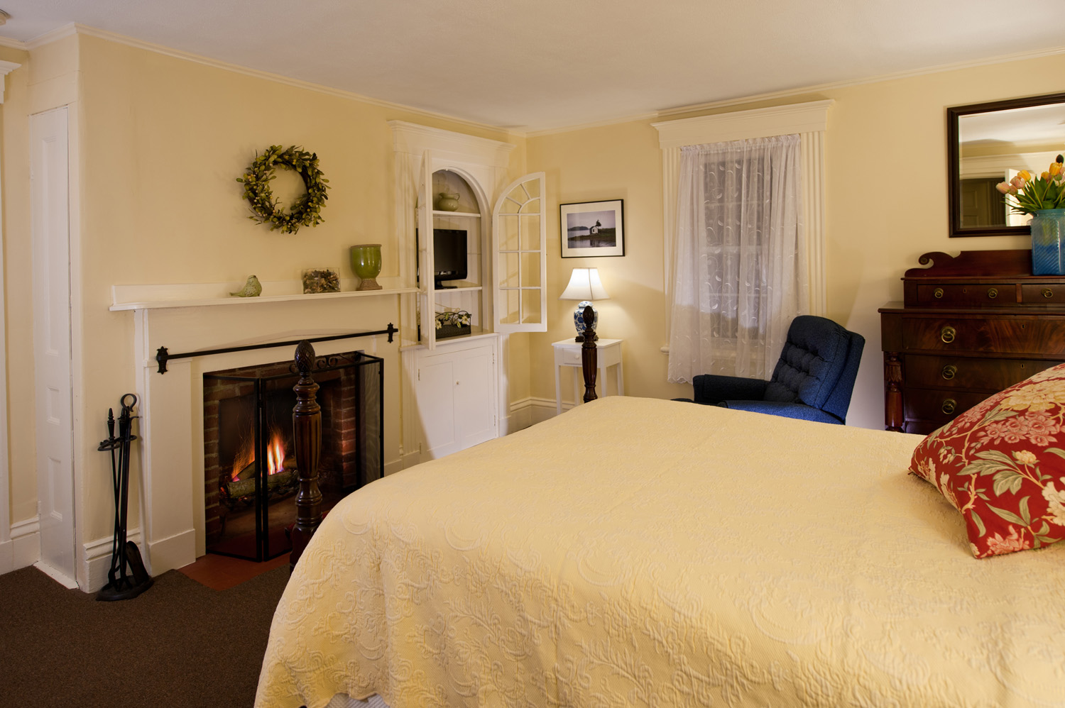 A yellow bedroom with bed with yellow bedspread and red floral pillow, a fireplace with log fire, and blue chair.