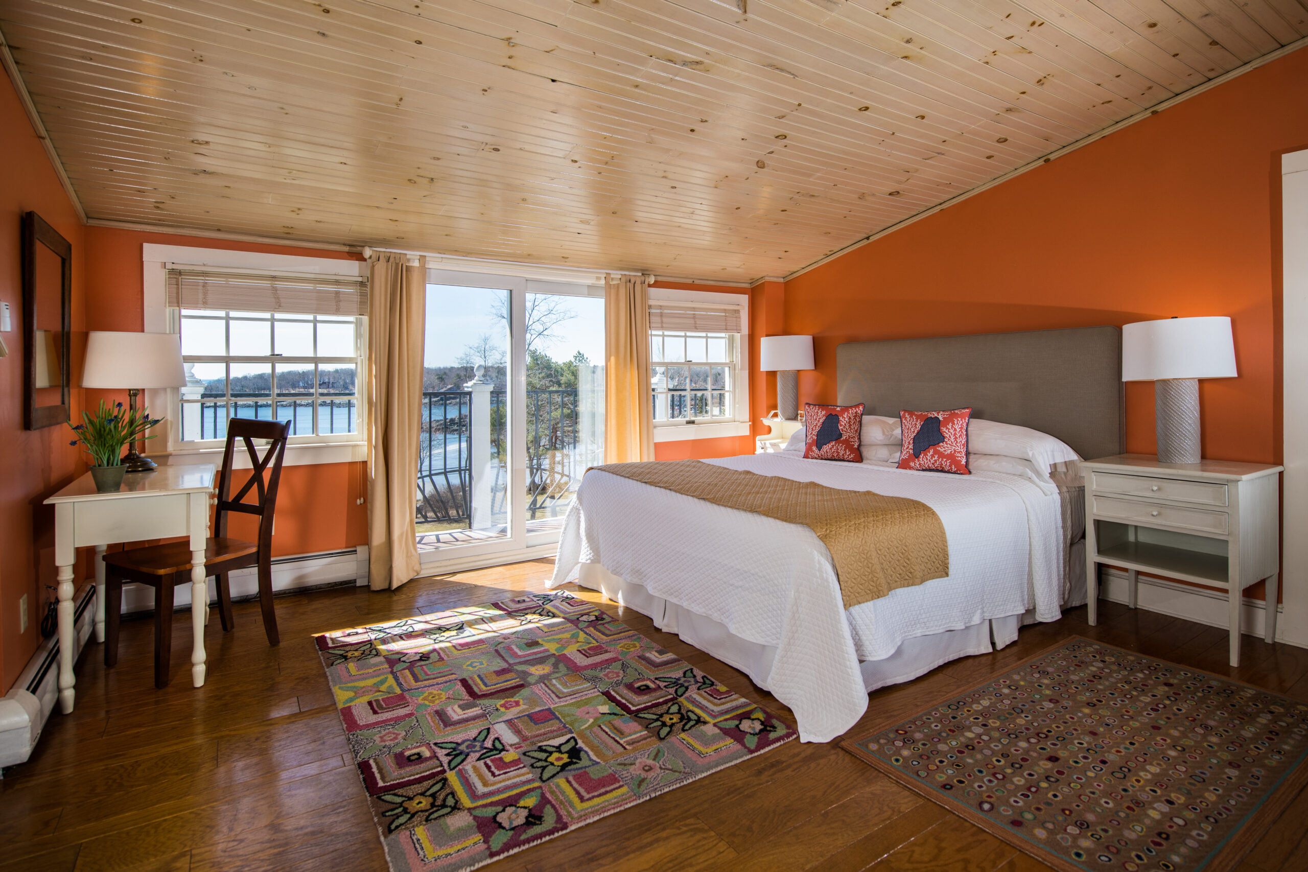 A coral bedroom with wooden floors, a pink, green, yelllow patterned rug, a desk, and king bed with a view through sliding doors of a balcony with ocean beyond.