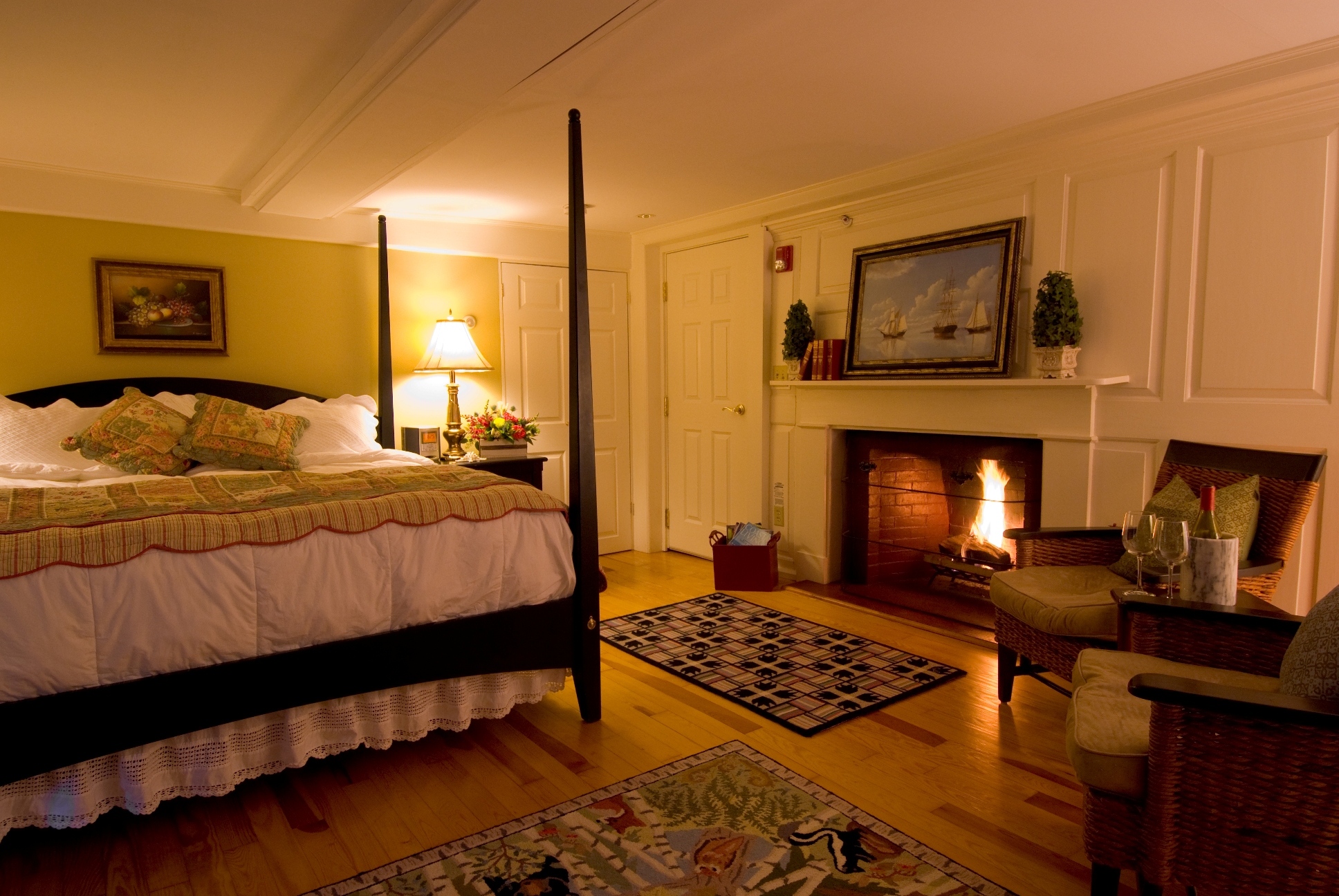 A four poster bed white linens, wood burning fireplace, and two rattan whicker chairs.